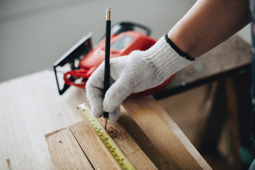 gloved hand using pencil and tape measure to measure piece of wood