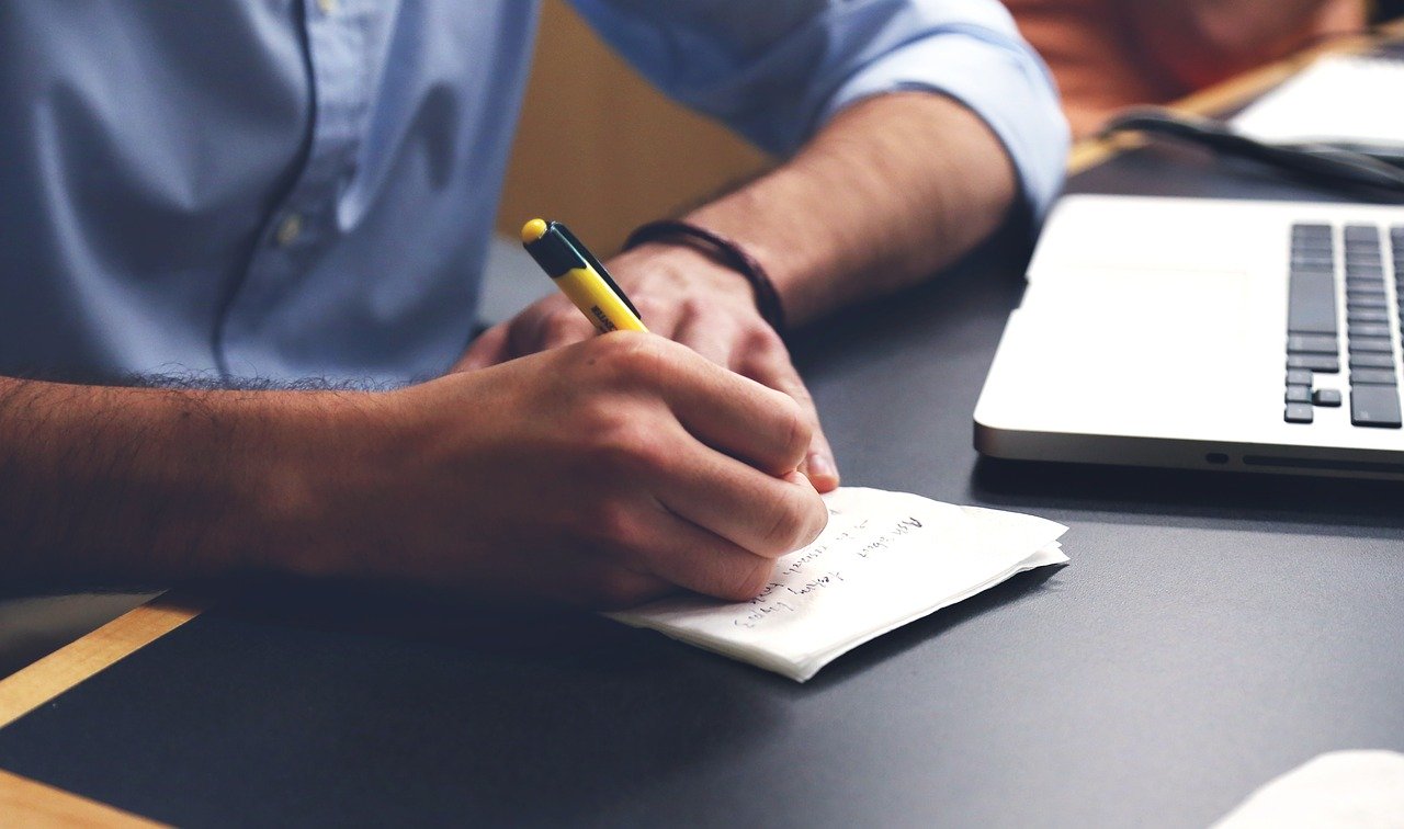 A man in a business shirt writing on a piece of paper in from of a computer