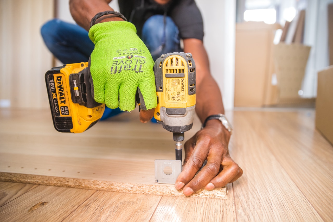 A worker with a green glove holding a yellow Dewalt hand drill drilling a screw