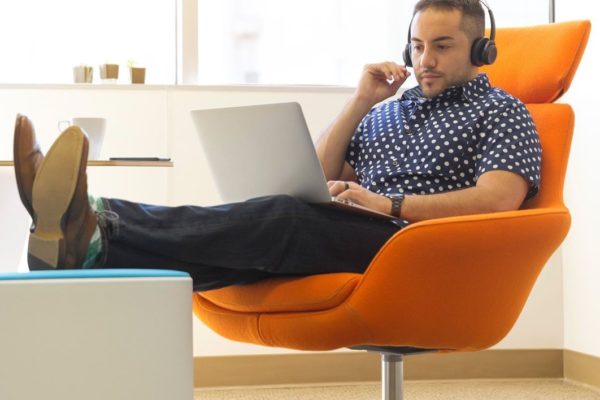 A man sitting in an orange chair wearing headphones and working on a laptop.