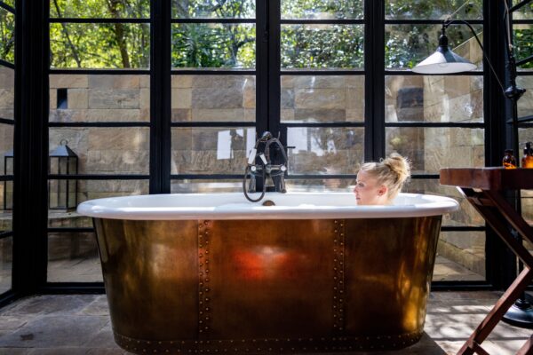 Woman sitting in a bathtub by large windows, enjoying natural light and relaxation.
